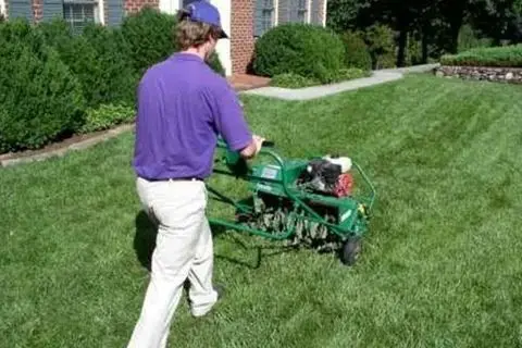 A person walks a commercial-grade aerating machine across a green lawn.
